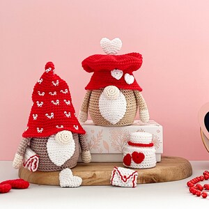 May include: Two crocheted gnomes with red and white hats and white beards. The gnomes are sitting on a wooden surface with a white box behind them. There are also two small crocheted hearts and a crocheted envelope on the surface.