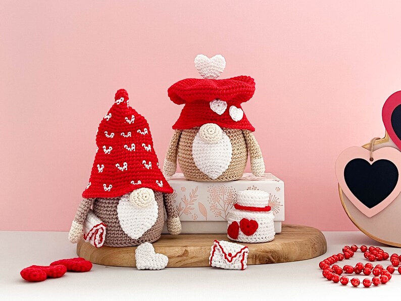 May include: Two crocheted gnomes with red and white hats and white beards. The gnomes are sitting on a wooden surface with a white box behind them. There are also two small crocheted hearts and a crocheted envelope on the surface.