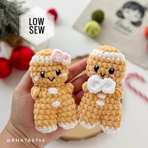 May include: Two small, tan, crocheted gingerbread figures with white icing details. One figure features a light pink bow on its head, while the other has a white bow at its neck. Both have black eyes, pink cheeks, and white accents on their feet and torso.