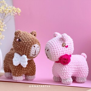May include: Two handmade crochet animal figurines. One is brown with a white bow tie, the other is pink with a pink flower collar. Both have gray accents and are set against a pink backdrop.