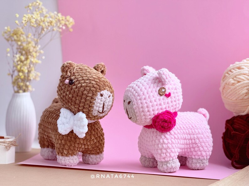 May include: Two handmade crochet animal figurines. One is brown with a white bow tie, the other is pink with a pink flower collar. Both have gray accents and are set against a pink backdrop.