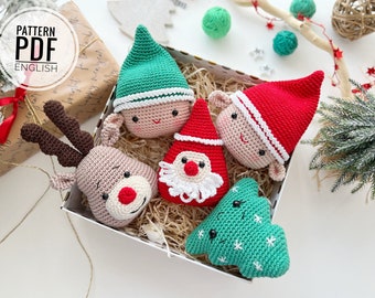 Puede incluir: Un conjunto de cinco adornos navideños de crochet en una caja blanca con papel triturado marrón. Los adornos incluyen un reno, un Papá Noel, un árbol de Navidad y dos duendes.