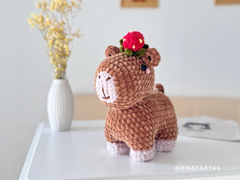 May include: A handmade brown crochet animal with a strawberry hat. The plush toy has a light pink face and feet. A small vase with yellow flowers is in the background.