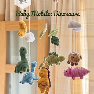 Può includere: Mobile per neonati fatto a mano con dinosauri all'uncinetto in vari colori, tra cui verde, blu, giallo e rosa. Il mobile include anche foglie, nuvole e una forma a mezzaluna all'uncinetto, sospesi a un telaio di legno. Il testo "Baby Mobile: Dinosaurs" è visibile.