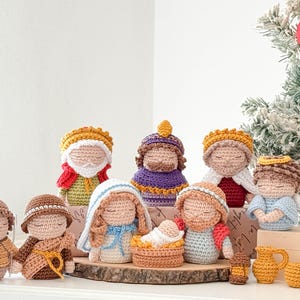 May include: A set of crocheted nativity figurines featuring various characters in colorful yarn outfits, including a central infant in a cradle, standing on a wooden base with small accessories like a pitcher and baskets arranged in the foreground.