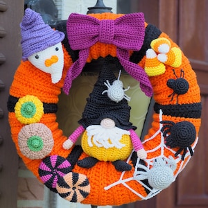 Puede incluir: Una corona de Halloween tejida a crochet con base naranja, rayas blancas y negras y varias decoraciones de temática de Halloween, incluyendo un gnomo, un fantasma, maíz dulce y arañas.