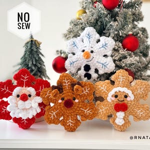 May include: Three plush crochet snowflake ornaments featuring character faces: a red Santa, a brown reindeer, and a white snowman. Each snowflake has embroidered white details and a soft texture. A white hexagon label reads NO SEW in black text.