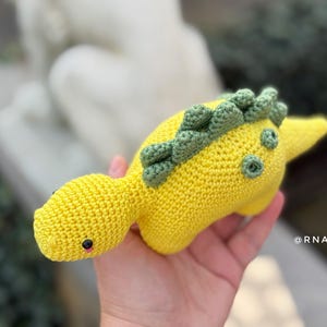 May include: A handmade, yellow crocheted dinosaur toy with green accents. The dinosaur has small black eyes and a pink mouth. The toy is held in a hand, showcasing its soft texture and detailed craftsmanship. The dinosaur's back features a row of green spikes.