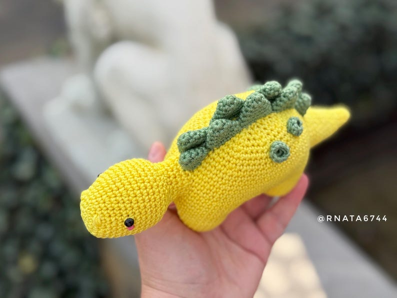 May include: A handmade, yellow crocheted dinosaur toy with green accents. The dinosaur has small black eyes and a pink mouth. The toy is held in a hand, showcasing its soft texture and detailed craftsmanship. The dinosaur's back features a row of green spikes.