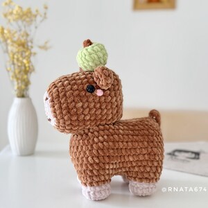 May include: A handmade brown crochet animal with a green apple on its head. The plush toy has a white muzzle, pink cheeks, and black eyes. The text "@RNATA6744" is visible on the right side. A unique, soft, and cuddly toy.