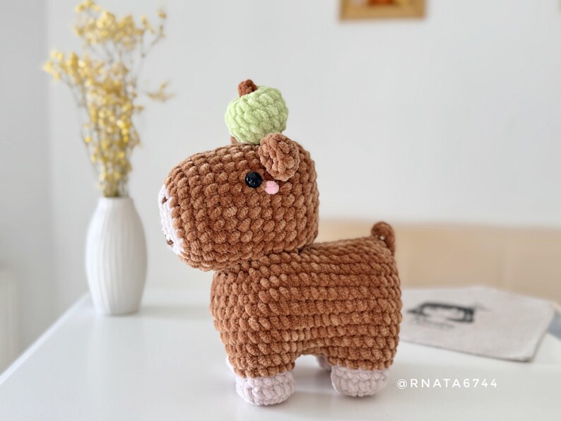 May include: A handmade brown crochet animal with a green apple on its head. The plush toy has a white muzzle, pink cheeks, and black eyes. The text "@RNATA6744" is visible on the right side. A unique, soft, and cuddly toy.
