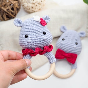 May include: Two handmade crochet baby rattles shaped like hippos. One hippo has a red bow and the other has a red ruffle collar and a white flower. The rattles are light grey with wooden rings.