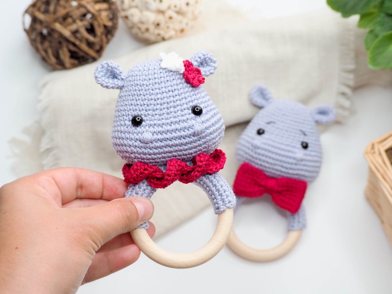 May include: Two handmade crochet baby rattles shaped like hippos. One hippo has a red bow and the other has a red ruffle collar and a white flower. The rattles are light grey with wooden rings.