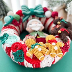 May include: A crocheted Christmas wreath with a red and white candy cane stripe pattern. The wreath is decorated with crocheted gingerbread people, a Santa Claus, a bow, and other festive ornaments.