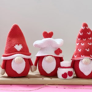 May include: Three crocheted gnomes in red and white with heart details. The gnomes are wearing hats and have white beards. One gnome is holding a red heart on a stick.
