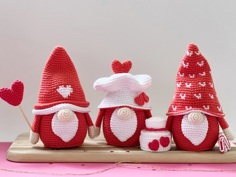 May include: Three crocheted gnomes in red and white with heart details. The gnomes are wearing hats and have white beards. One gnome is holding a red heart on a stick.