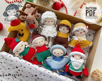 Puede incluir: Una colección de adornos navideños de crochet hechos a mano en una caja. Los adornos incluyen un reno, una luna, un muñeco de nieve, una niña, un niño, un Papá Noel y un duende. Los adornos son todos de diferentes colores y tienen diferentes expresiones faciales. La caja está forrada con papel blanco con un patrón de árbol de Navidad.