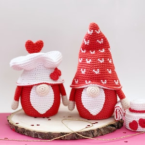 May include: Two crocheted gnomes in red and white with heart details. The gnomes are standing on a wooden slice with red hearts scattered around them.