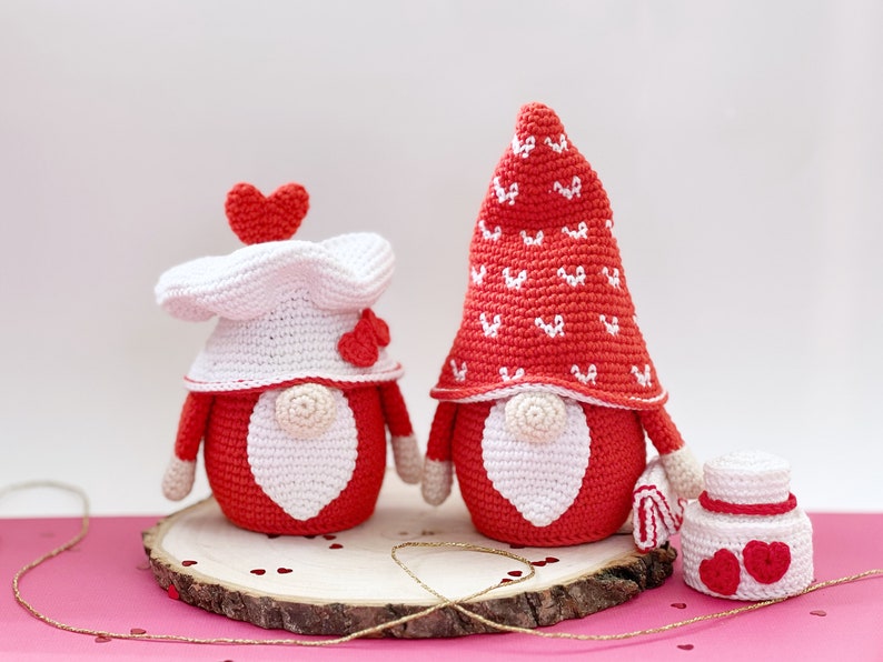 May include: Two crocheted gnomes in red and white with heart details. The gnomes are standing on a wooden slice with red hearts scattered around them.