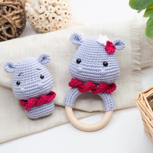 May include: Two crocheted hippo baby toys. One is a rattle with a wooden ring, and the other is a small hippo head. Both are light blue with red accents and black eyes. The rattle has a red scarf around its neck.