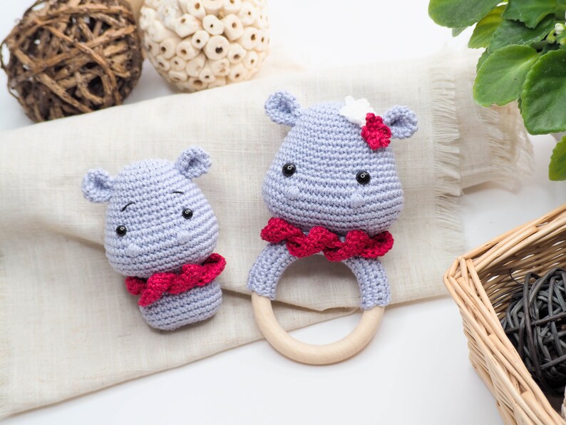 May include: Two crocheted hippo baby toys. One is a rattle with a wooden ring, and the other is a small hippo head. Both are light blue with red accents and black eyes. The rattle has a red scarf around its neck.