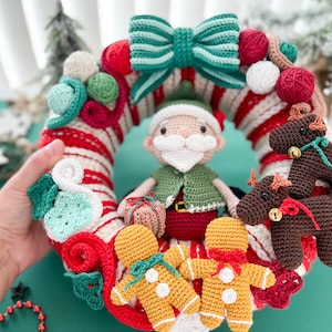 May include: A crocheted Christmas wreath with a red and white candy cane design. The wreath is decorated with a green and white bow, a green elf holding a gift, two gingerbread men, and two brown reindeer.
