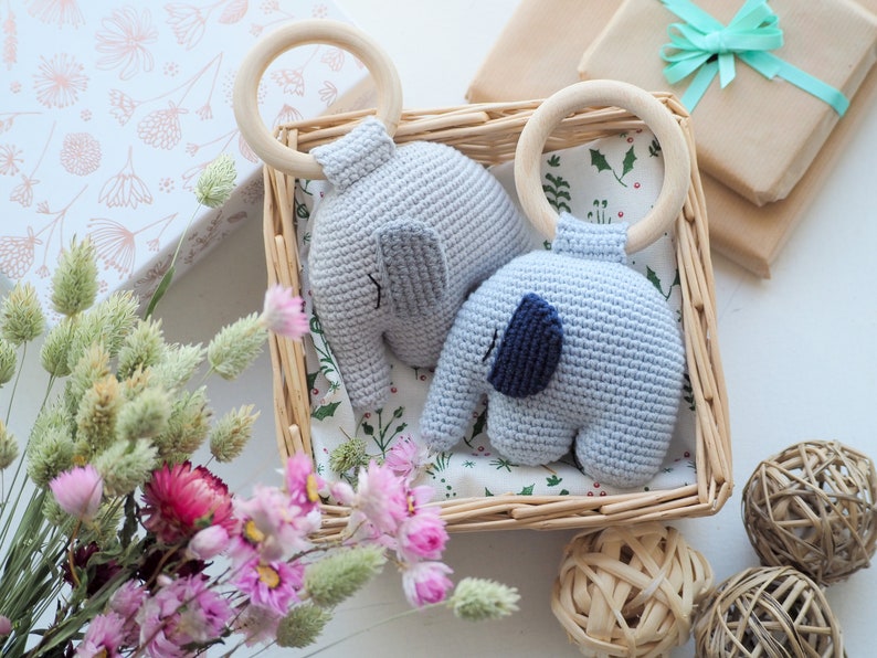 Puede incluir: Dos sonajeros de beb&eacute; de elefante de crochet con anillos de madera en una cesta de mimbre. Los elefantes son de color azul claro y gris con detalles bordados. La cesta est&aacute; forrada con una tela blanca con un patr&oacute;n floral.