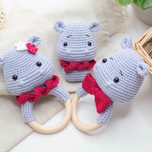 May include: Three crocheted hippo baby rattles in light blue with red accents. Two rattles have wooden rings and red bows, while the third has a red scarf. One hippo has a white flower. The rattles are handmade.