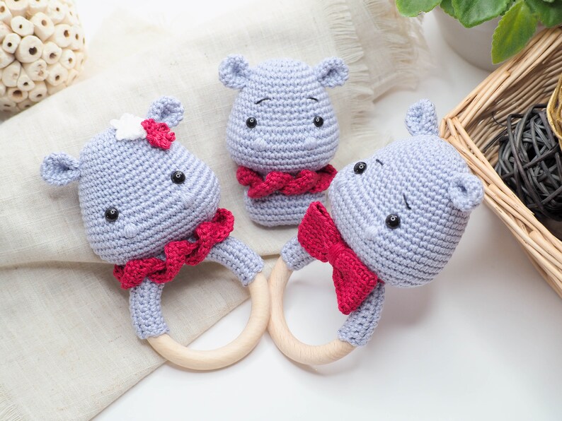 May include: Three crocheted hippo baby rattles in light blue with red accents. Two rattles have wooden rings and red bows, while the third has a red scarf. One hippo has a white flower. The rattles are handmade.