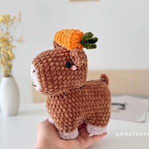 May include: A handmade brown crochet animal with a carrot on its head. The plush toy has a white face, pink accents, and small black eyes. The carrot is orange with green leaves. The toy is held in a person's hand.
