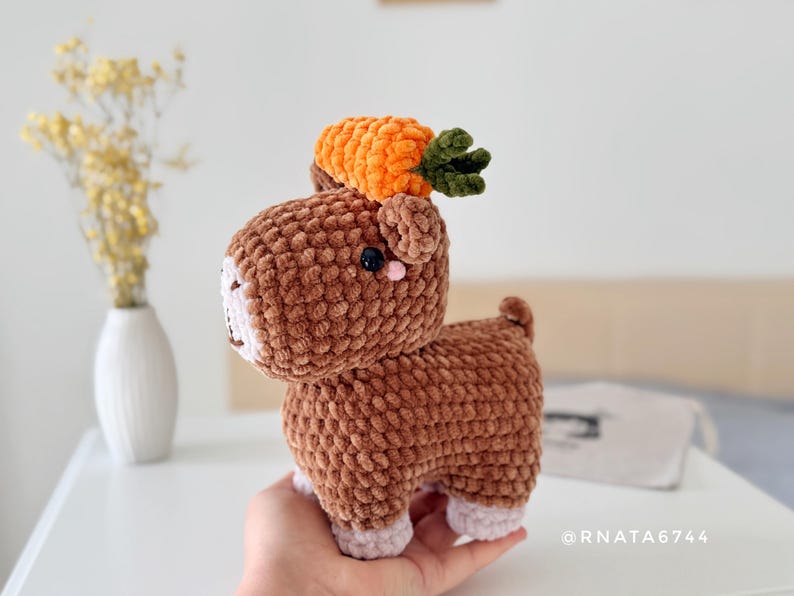 May include: A handmade brown crochet animal with a carrot on its head. The plush toy has a white face, pink accents, and small black eyes. The carrot is orange with green leaves. The toy is held in a person's hand.