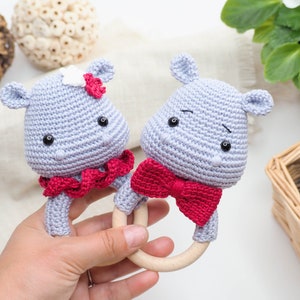 May include: Two handmade crochet hippo baby rattles. The hippos are light grey with black eyes and red accents. One hippo has a red bow tie, and the other has a red and white flower. The rattles have wooden ring handles.
