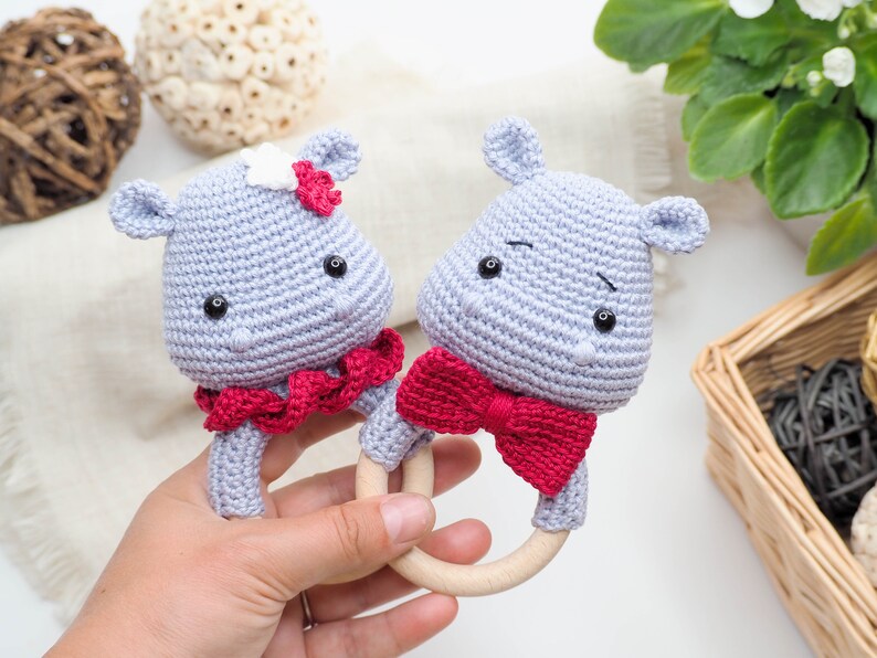 May include: Two handmade crochet hippo baby rattles. The hippos are light grey with black eyes and red accents. One hippo has a red bow tie, and the other has a red and white flower. The rattles have wooden ring handles.