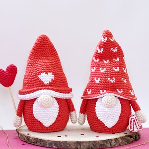May include: Two crocheted gnomes with red hats and white beards. One gnome's hat has a white heart design, and the other has a white heart pattern. The gnomes are holding a red heart.