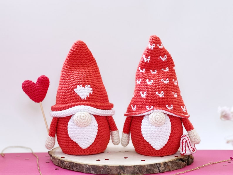 May include: Two crocheted gnomes with red hats and white beards. One gnome's hat has a white heart design, and the other has a white heart pattern. The gnomes are holding a red heart.