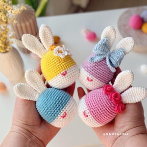 Pode incluir: Quatro enfeites de coelhinhos da Páscoa feitos à mão em crochê nas cores amarelo, roxo, azul e rosa. Cada coelhinho tem orelhas brancas, um rosto branco e um pequeno sorriso bordado. O coelhinho amarelo tem uma flor, o roxo um laço e o rosa uma rosa.