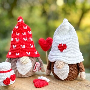 May include: Two crocheted gnomes with red and white hats and brown faces. The gnome on the left has a red hat with white hearts and is holding a white cake with red hearts. The gnome on the right has a white hat with a red heart and is holding a red heart on a stick.