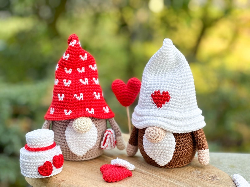 May include: Two crocheted gnomes with red and white hats and brown faces. The gnome on the left has a red hat with white hearts and is holding a white cake with red hearts. The gnome on the right has a white hat with a red heart and is holding a red heart on a stick.