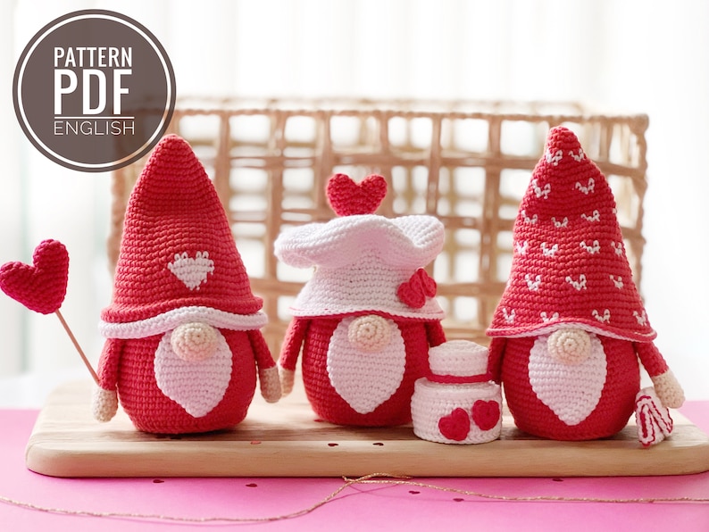 May include: Three crocheted gnomes with red hats and white beards. The gnome on the left is holding a red heart on a stick. The gnome in the middle has a white hat with a red heart on top. The gnome on the right has a red hat with white hearts on it. The gnomes are sitting on a wooden surface.  PATTERN PDF ENGLISH