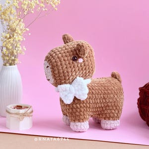 May include: A handmade brown crochet goat with a white bib and pink accents. The amigurumi animal has a textured appearance and is standing on a pink surface. A white vase with yellow flowers and yarn balls are in the background.