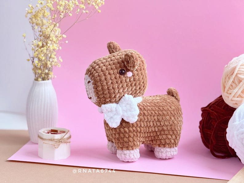 May include: A handmade brown crochet goat with a white bib and pink accents. The amigurumi animal has a textured appearance and is standing on a pink surface. A white vase with yellow flowers and yarn balls are in the background.