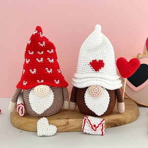 May include: Two crocheted gnomes with red and white hearts on their hats. The gnome on the left has a red hat with white hearts and the gnome on the right has a white hat with a red heart. Both gnomes have brown bodies and white beards.