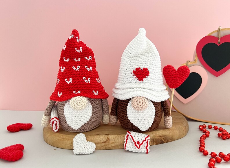 May include: Two crocheted gnomes with red and white hearts on their hats. The gnome on the left has a red hat with white hearts and the gnome on the right has a white hat with a red heart. Both gnomes have brown bodies and white beards.