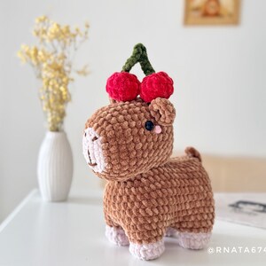 May include: A handmade brown crochet animal with a cherry on its head. The plush toy has a light pink face and feet, with a dark blue eye. The cherries are red with a green stem. The toy is on a white surface.
