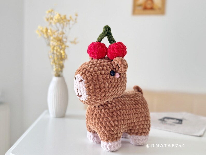 May include: A handmade brown crochet animal with a cherry on its head. The plush toy has a light pink face and feet, with a dark blue eye. The cherries are red with a green stem. The toy is on a white surface.