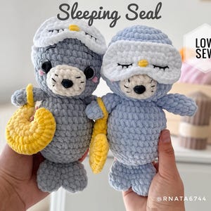 Könnte beinhalten: Zwei gehäkelte Seehund-Plüschtiere, eines grau und eines blau, tragen jeweils eine weiße Schlafmaske mit gelbem Akzent. Sie halten gelbe Accessoires. Der Text "Sleeping Seal" steht darüber.