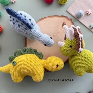 May include: Three handmade crochet dinosaur toys in blue, yellow, and green. The dinosaurs are on a wooden slice with yarn and decorative elements in the background. The toys have black eyes and are made with soft yarn.