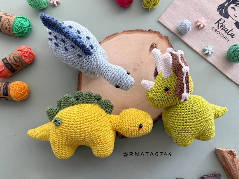 May include: Three handmade crochet dinosaur toys in blue, yellow, and green. The dinosaurs are on a wooden slice with yarn and decorative elements in the background. The toys have black eyes and are made with soft yarn.