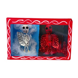 May include: A red wooden frame with a glass front, containing two small skeleton figures. The skeleton on the left is white and the skeleton on the right is red. The background behind the skeletons is painted blue and red.