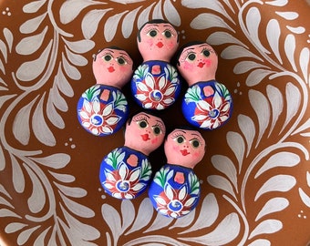 May include: Seven small, hand-painted ceramic dolls with blue and white floral patterns and pink faces. The dolls are arranged in a circle on a terracotta plate with a white floral design.